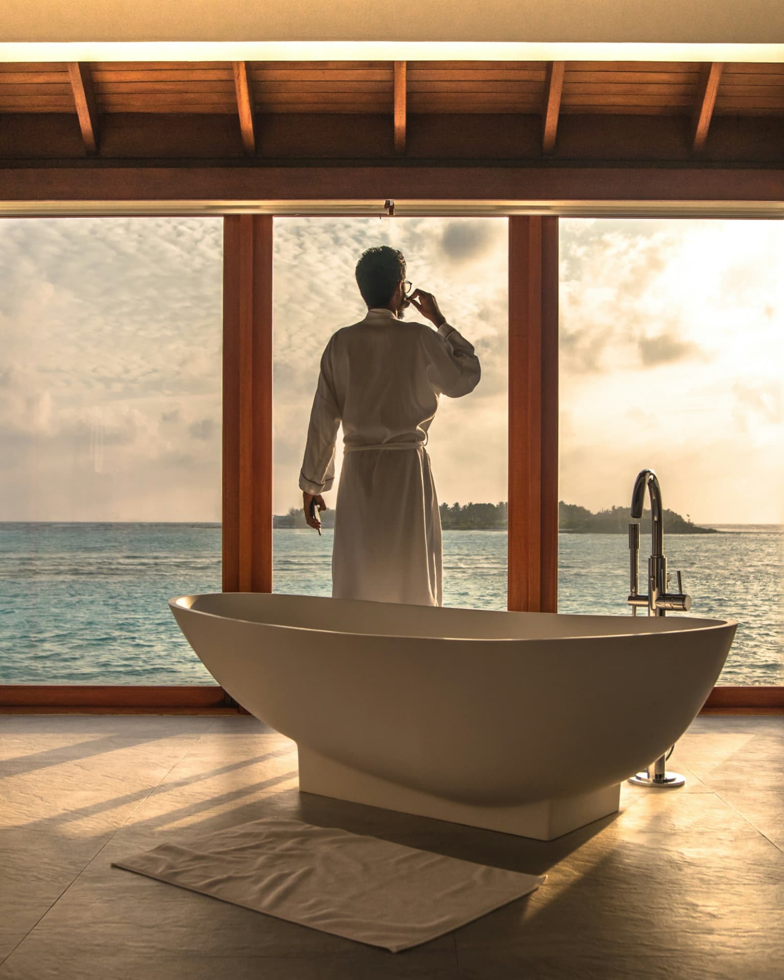 Traveler in a robe looking out over the ocean from a villa bathroom